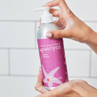 Hand holding a bottle of 'plane products' shampoo with cherry almond scent against a tiled wall background.