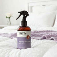 Bottle of 'Blissful Dreams' pillow spray on a bed with white bedding and a vase in the background.