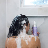 Person with soapy hair holding a bottle of body wash in a bathroom.