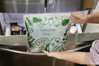 Person holding a smoothie pack with green vegetable illustrations in a kitchen setting
