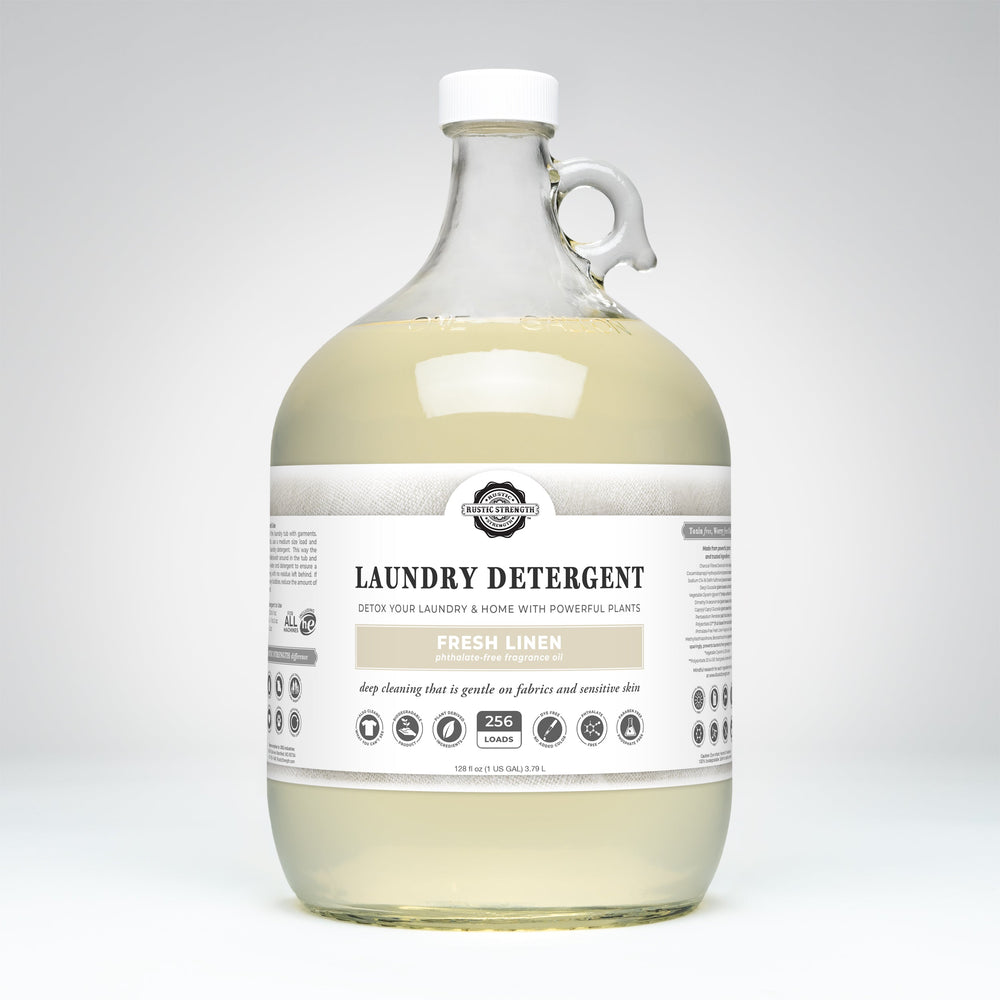 A large clear plastic jug of laundry detergent with a white cap and handle, filled with pale yellow liquid. The front label reads 'The Laundry Company' and 'Laundry Detergent' in 'Fresh Linen'.