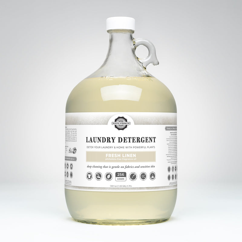 A large clear plastic jug of laundry detergent with a white cap and handle, filled with pale yellow liquid. The front label reads 'The Laundry Company' and 'Laundry Detergent' in 'Fresh Linen'.