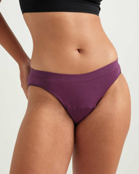 A close-up of a model wearing a pair of purple high-cut bikini bottoms, showing the waist, hips, and upper thighs against a plain light background.