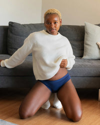 Person wearing a white sweater and blue underwear kneeling on a wooden floor with a gray couch in the background.