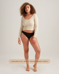 Woman wearing a beige long-sleeve top and black underwear on a plain background