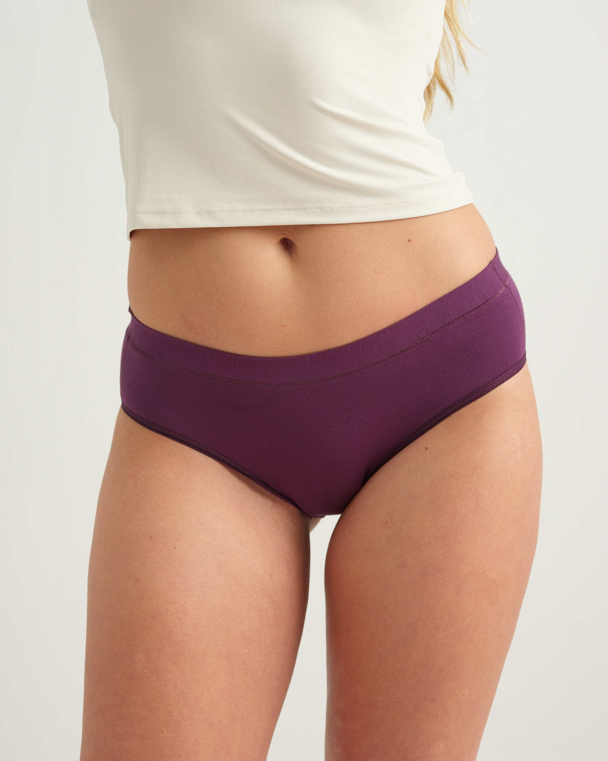 A close-up of a model wearing a pair of solid purple high-waisted briefs, paired with a white cropped tank top, against a light gray studio background. Main image
