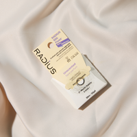 A single Radius Silk 11og eyeglass lens on its retail card, placed on draped white satin fabric. The packaging is cream and white with purple accents, and the lens itself is clear with a subtle blue reflection.