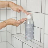 Person applying shampoo to hands with a bottle of shampoo in the foreground, against a tiled shower wall.