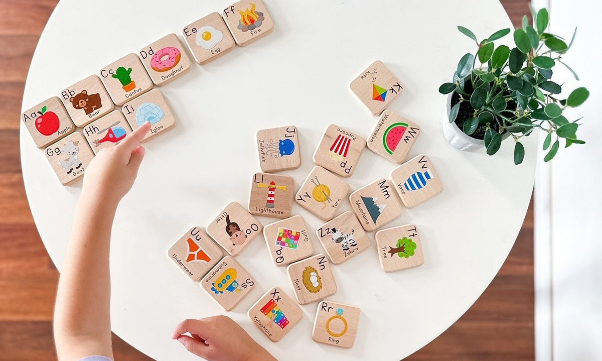 A top-down view of a child's hands interacting with a set of wooden alphabet and basic learning blocks arranged on a white round table next to a potted plant. Main image